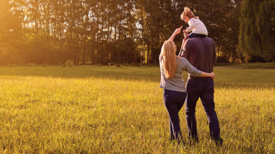 Husband and wife walk through wooded area with young daughter on father's shoulders